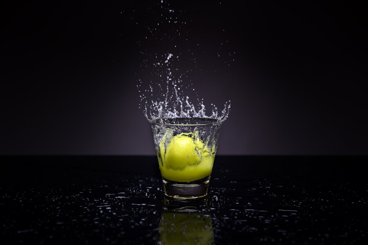 A lemon making a splash in a glass of water against a dark background.